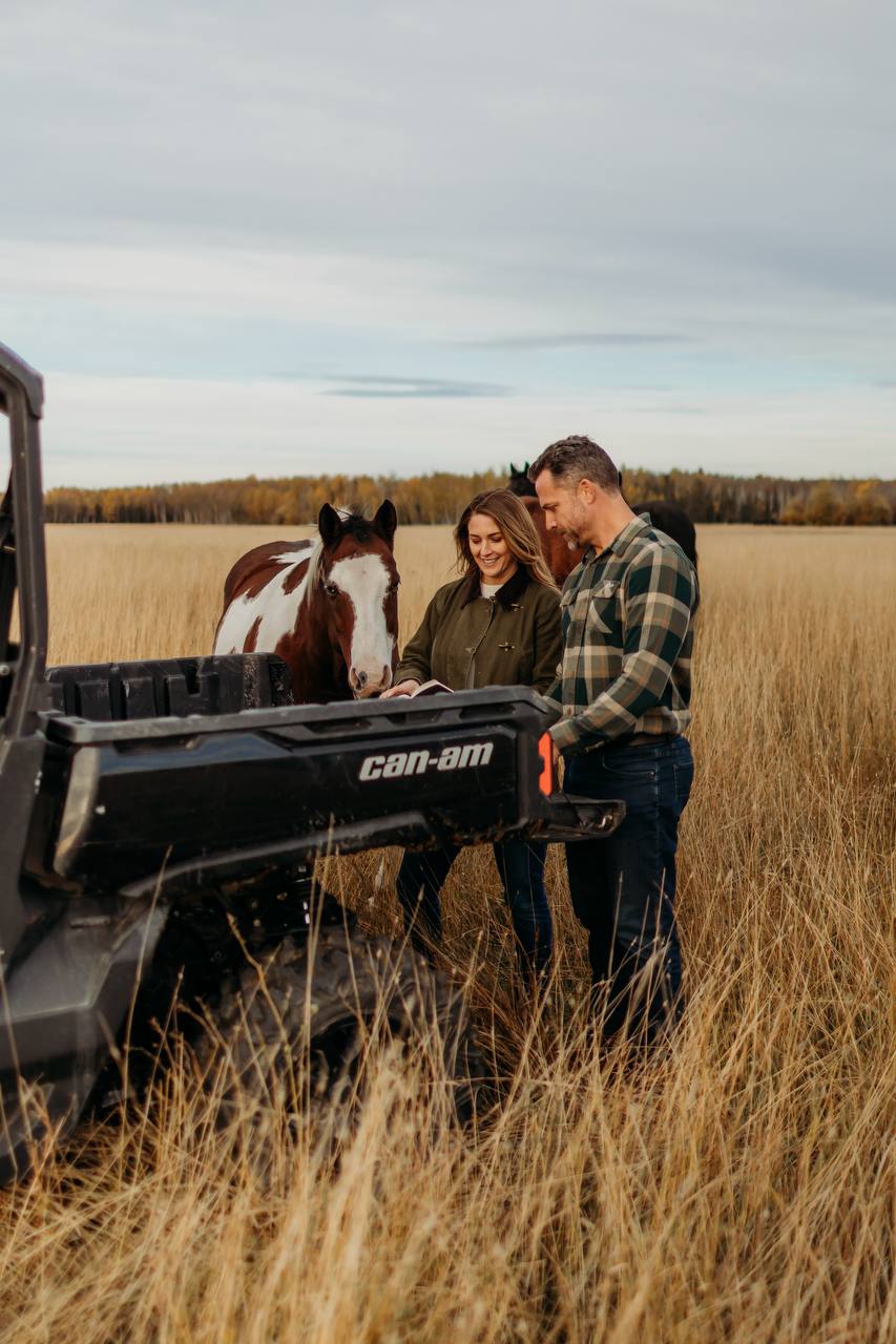 A woman and man reviewing paperwork beside a utility vehicle and horses in a grassy pasture.
