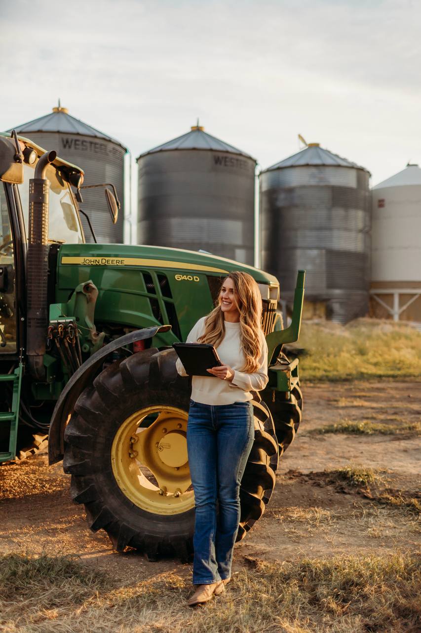 A woman holding a tablet beside a green tractor, with grain bins behind her.