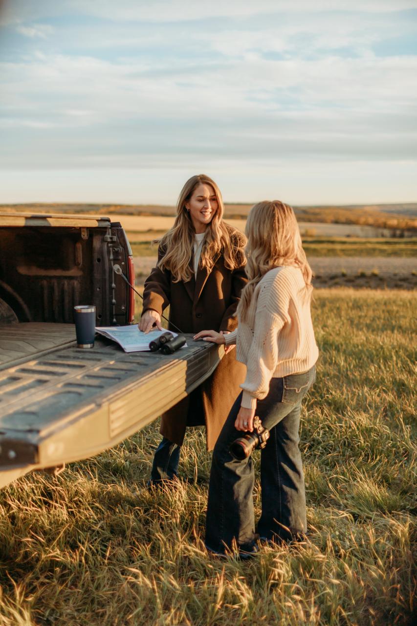 Two women reviewing a map on a pickup truck tailgate overlooking open farmland.
